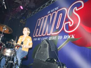 Drummer performing on stage in front of a vibrant backdrop featuring the band name "HINDS" and the phrase "AND WE CAME HERE TO ROCK," emphasizing promotional stage scrims and band banners.