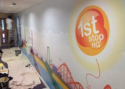 Wall mural featuring vibrant graphics of a cityscape and the text "1st stop HQ," with a person painting on a ladder, showcasing custom wallpaper installation for office environments.