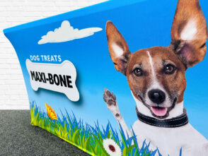Printed tablecloth featuring a smiling dog with a collar, promoting "Maxi-Bone" dog treats, set against a vibrant blue background with grass and flowers, suitable for exhibition displays.