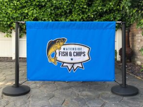 Cafe barrier featuring a blue printed banner for "Waterside Fish & Chips," supported by stainless steel posts and heavyweight bases, set in an outdoor environment.
