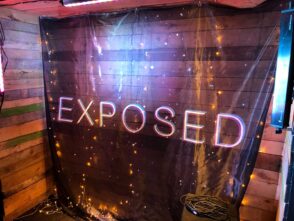 Backdrop featuring the word "EXPOSED" in illuminated letters, surrounded by decorative lights, set against a wooden wall, relevant to band backdrop printing options.